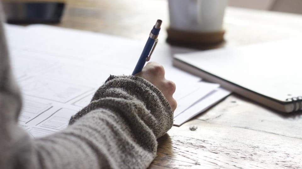 A hand writing notes with a pen on printed sheets beside a laptop and a coffee cup.